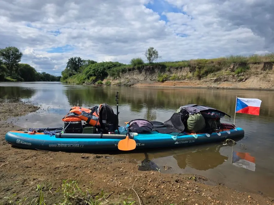 Nafouknutý paddleboard naložený výbavou na břehu řeky, s českou vlajkou
